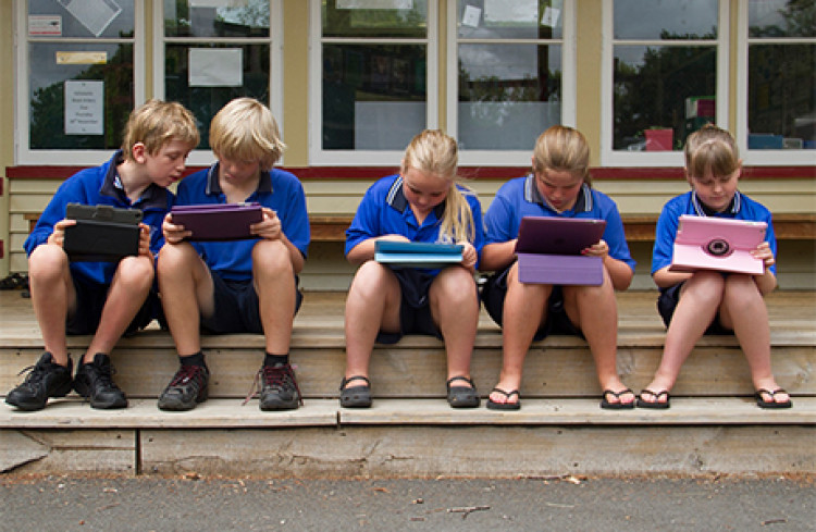 Five primary school aged children in school uniforms sitting on a step each working on electronic tablets.